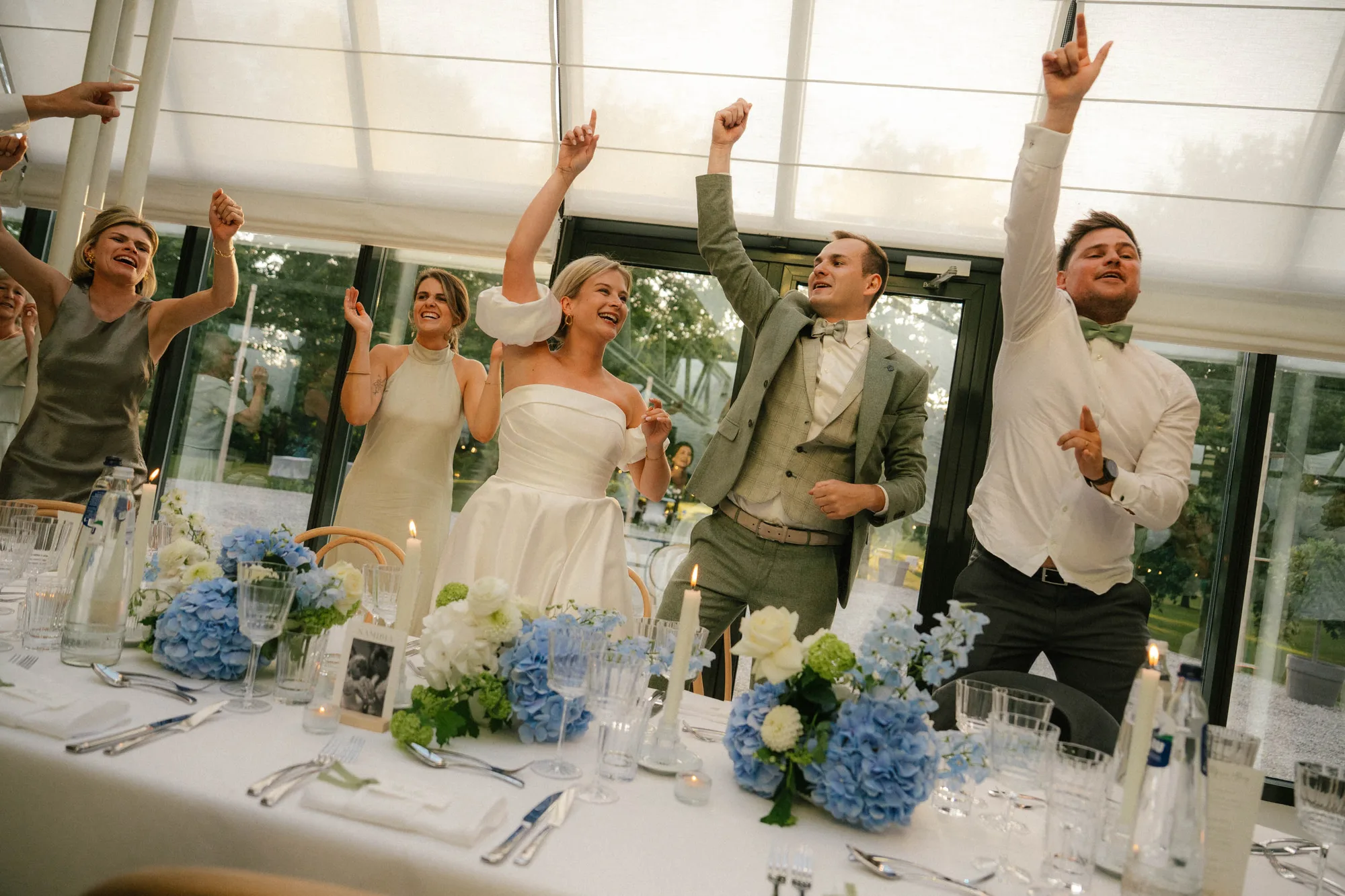 Wedding party with bride, groom, and guests dancing joyfully near a table decorated with blue and white flowers and candles.