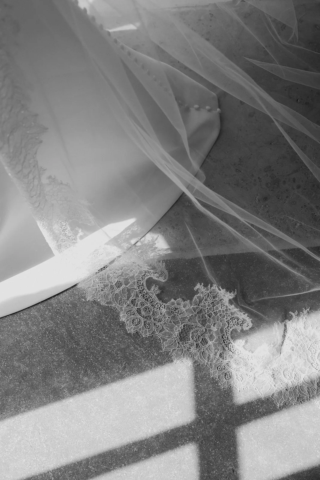 Close-up of a wedding dress train with lace edges and fabric-covered buttons on the floor with sunlight casting shadows.