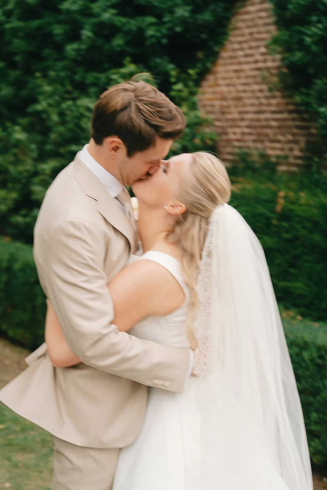 Bride in white dress and veil kissing groom in beige suit outdoors with greenery and brick wall background.