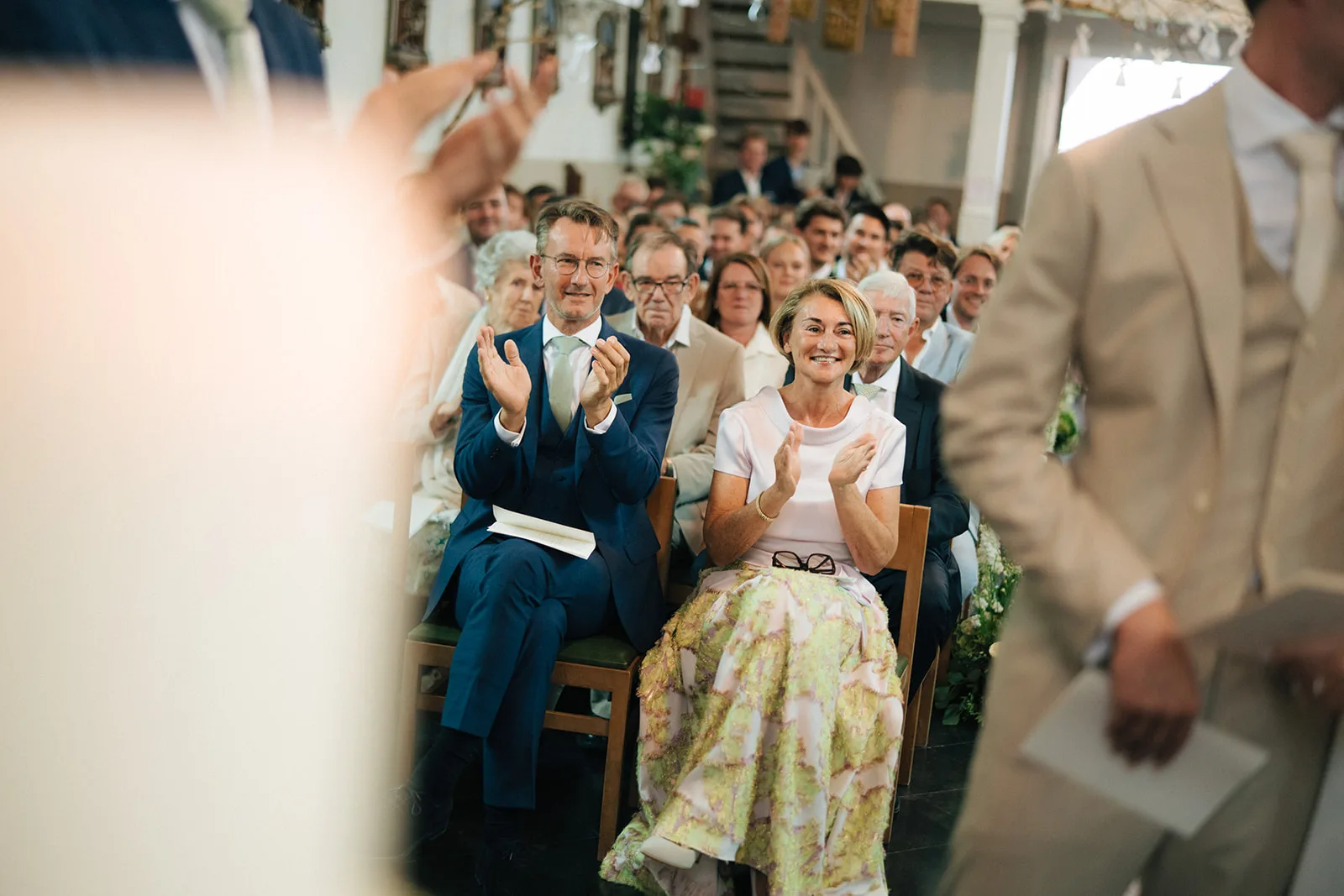 Guests in formal attire clapping and smiling while seated at a wedding ceremony.
