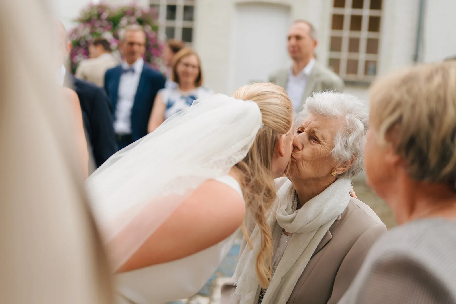 Bride in wedding dress and veil kissing an elderly woman wearing a scarf and coat, with blurred wedding guests in the background.