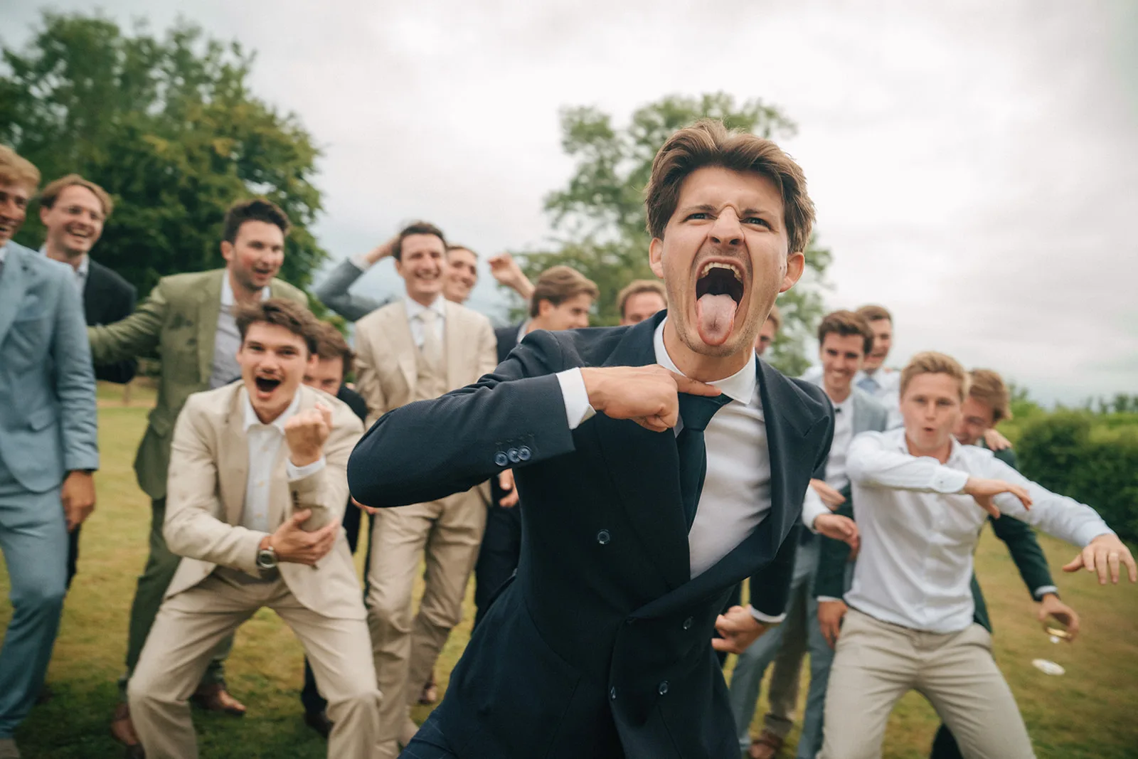 Group of men in suits and dress shirts outdoors, with one man in front making an energetic facial expression and gesture.