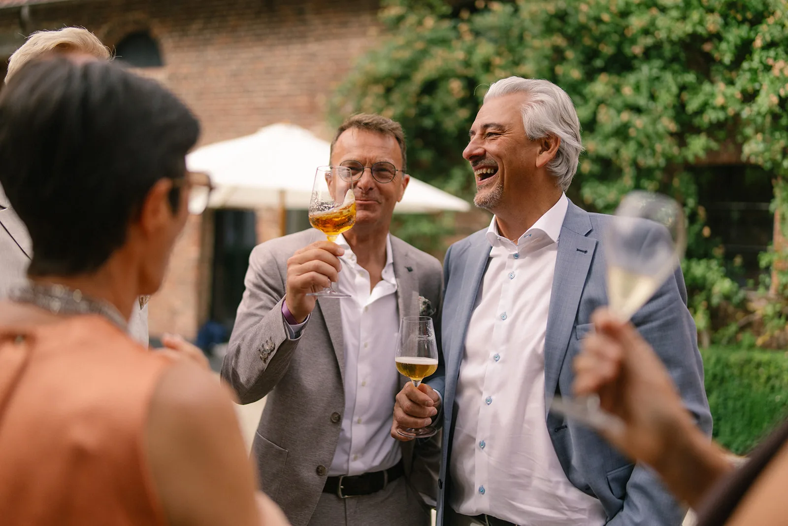 Two men in suits laughing and holding glasses of beer while socializing outdoors with other people.