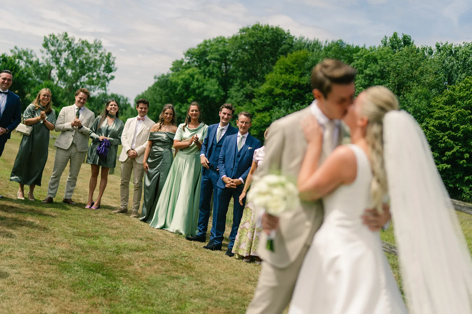 Bride and groom kissing in the foreground with a line of wedding guests dressed formally clapping and smiling in the background on a grassy lawn.