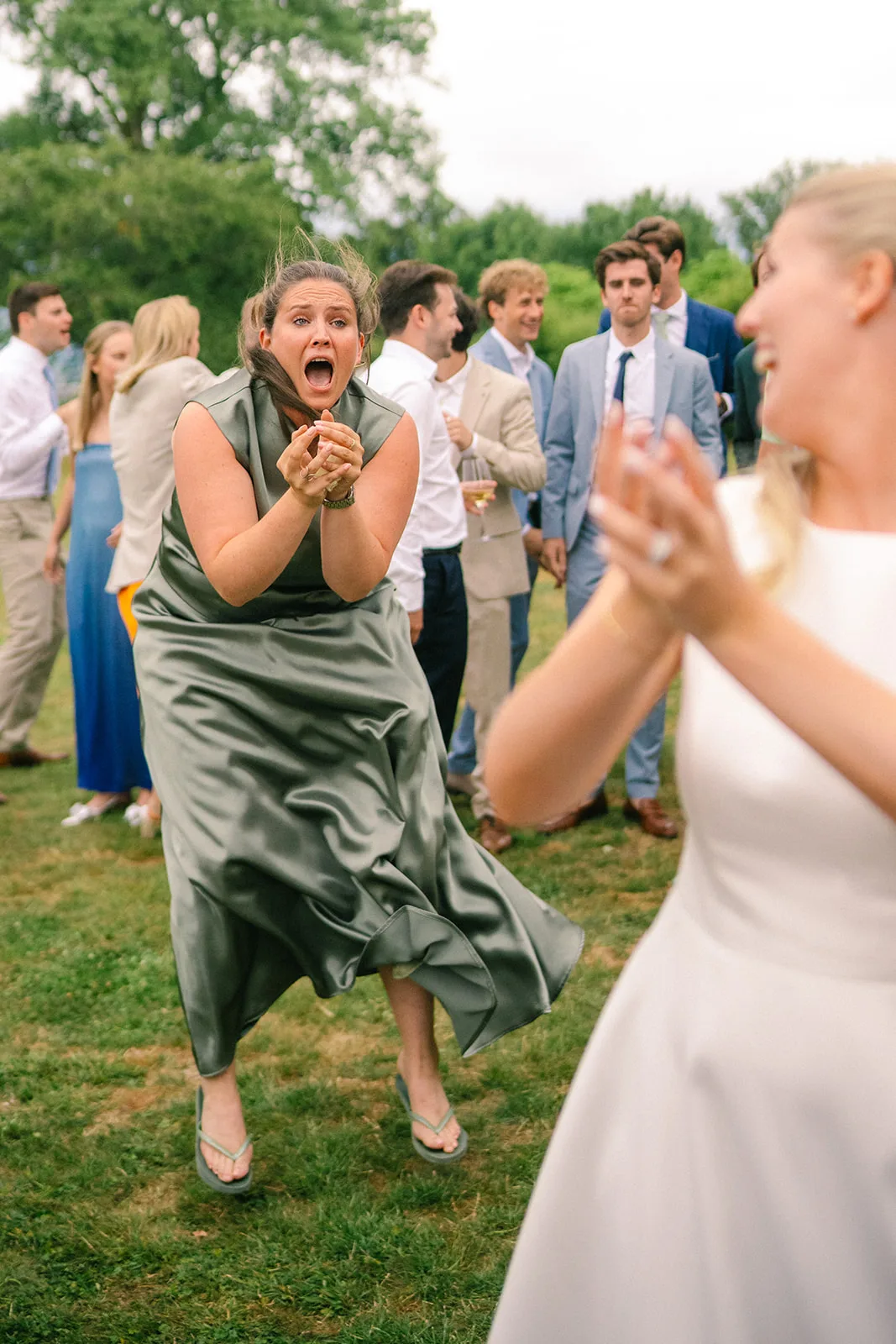Woman in a green dress excitedly reaching out with hands clasped outdoors while others, including a blurred bride in white, stand in the background on grass.