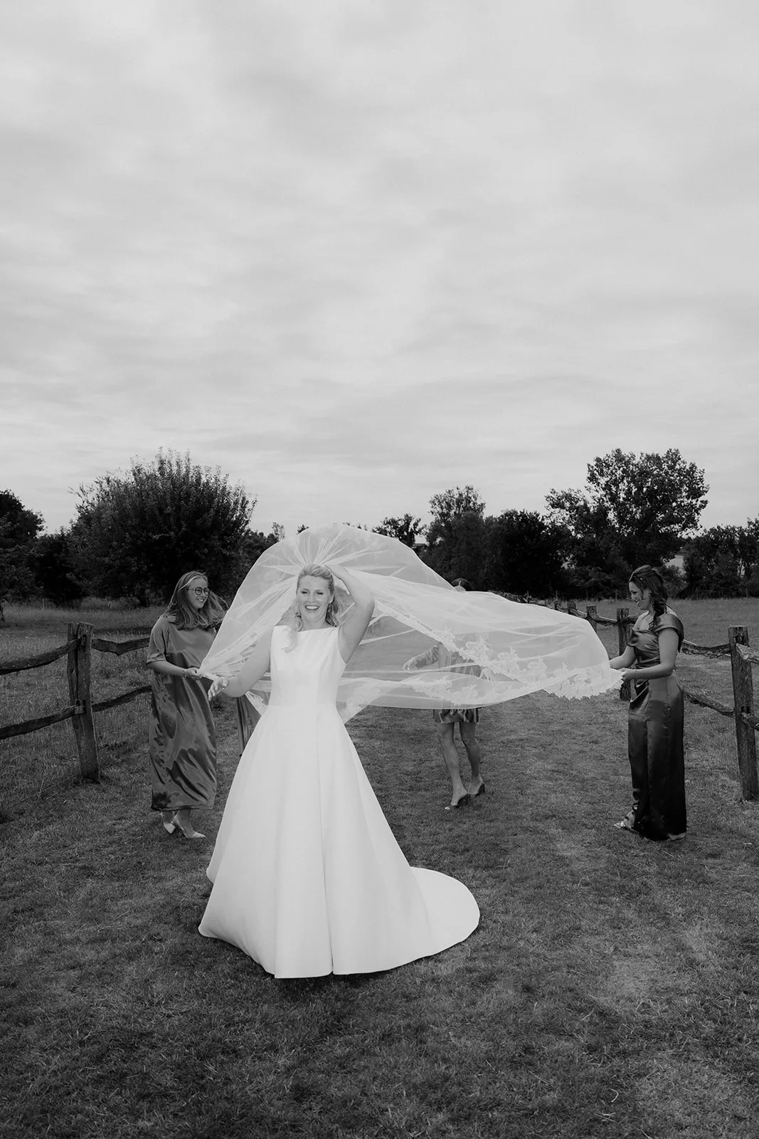 Bride in a white wedding dress with a long flowing veil held by two women in a field with wooden fences.