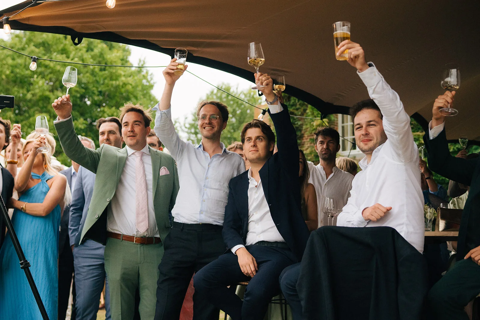 Group of men raising glasses in a toast at an outdoor event under a tent.