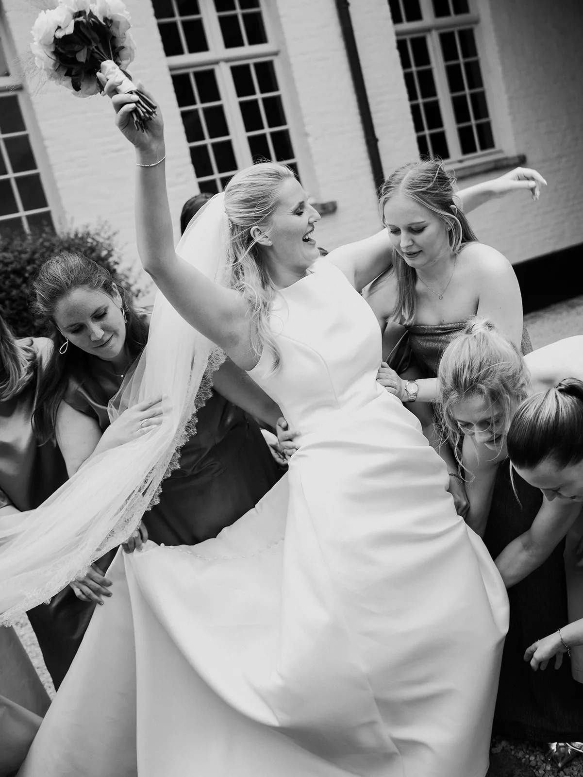 Bride smiling joyfully and raising bouquet while bridesmaids help adjust her wedding dress and veil outdoors.