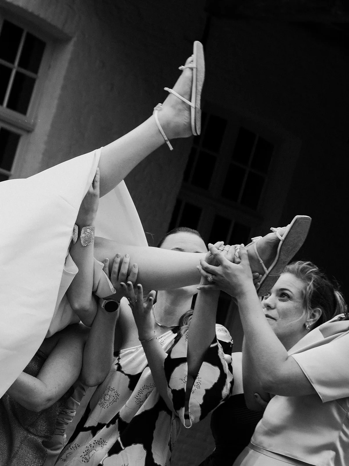 Several people lifting a person in a white dress wearing sandals into the air during a celebration.