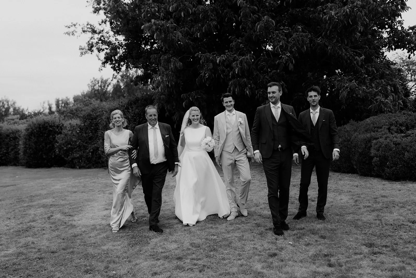 Black and white photo of a bride and groom walking on grass with four family members in formal attire, smiling.