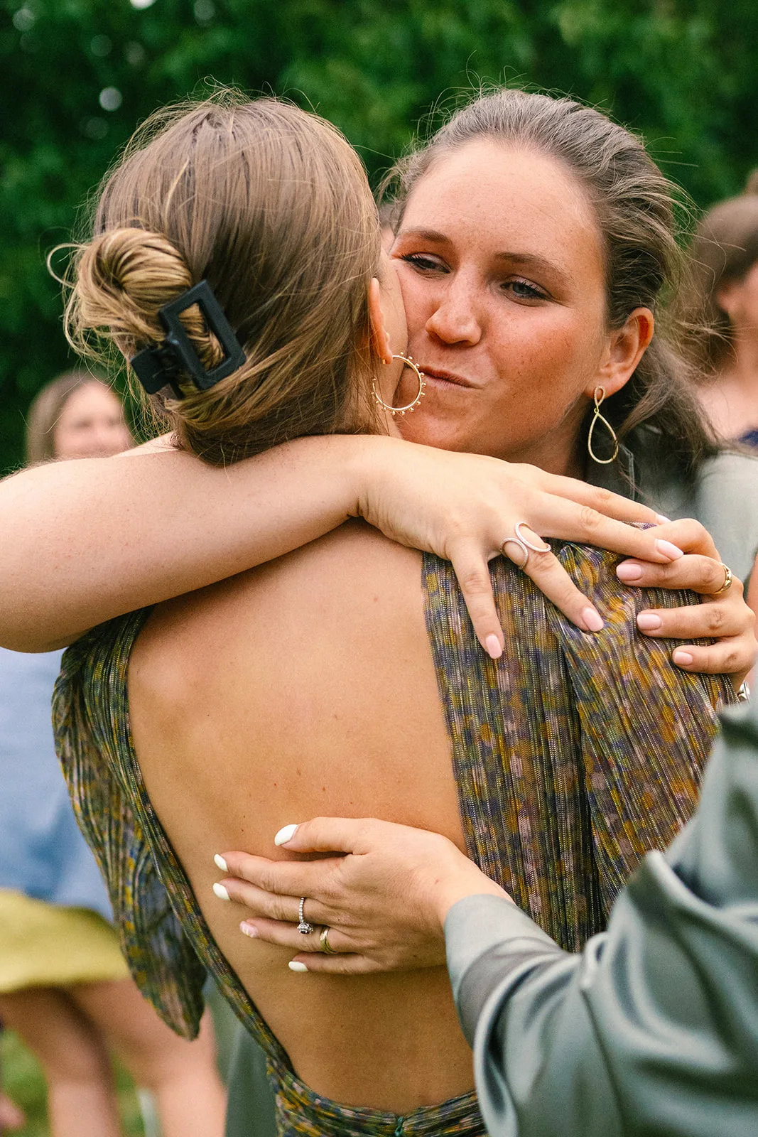 Two women embracing outdoors, one with an open-back dress and the other with hoop earrings and loose hair, sharing a close moment.