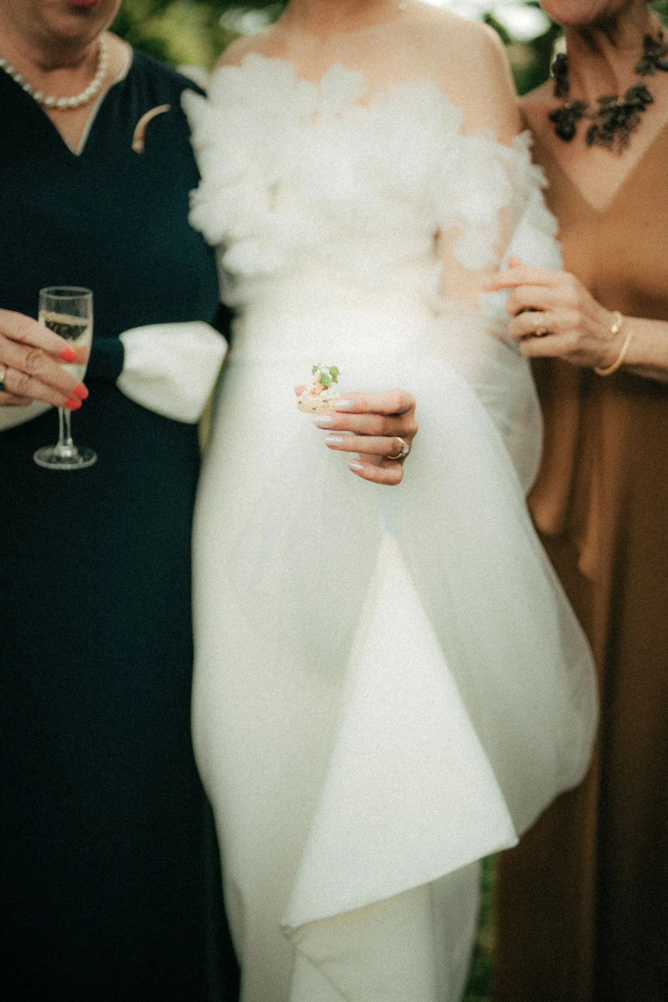 Close-up of three women holding a drink and an appetizer, one wearing a white off-shoulder dress.
