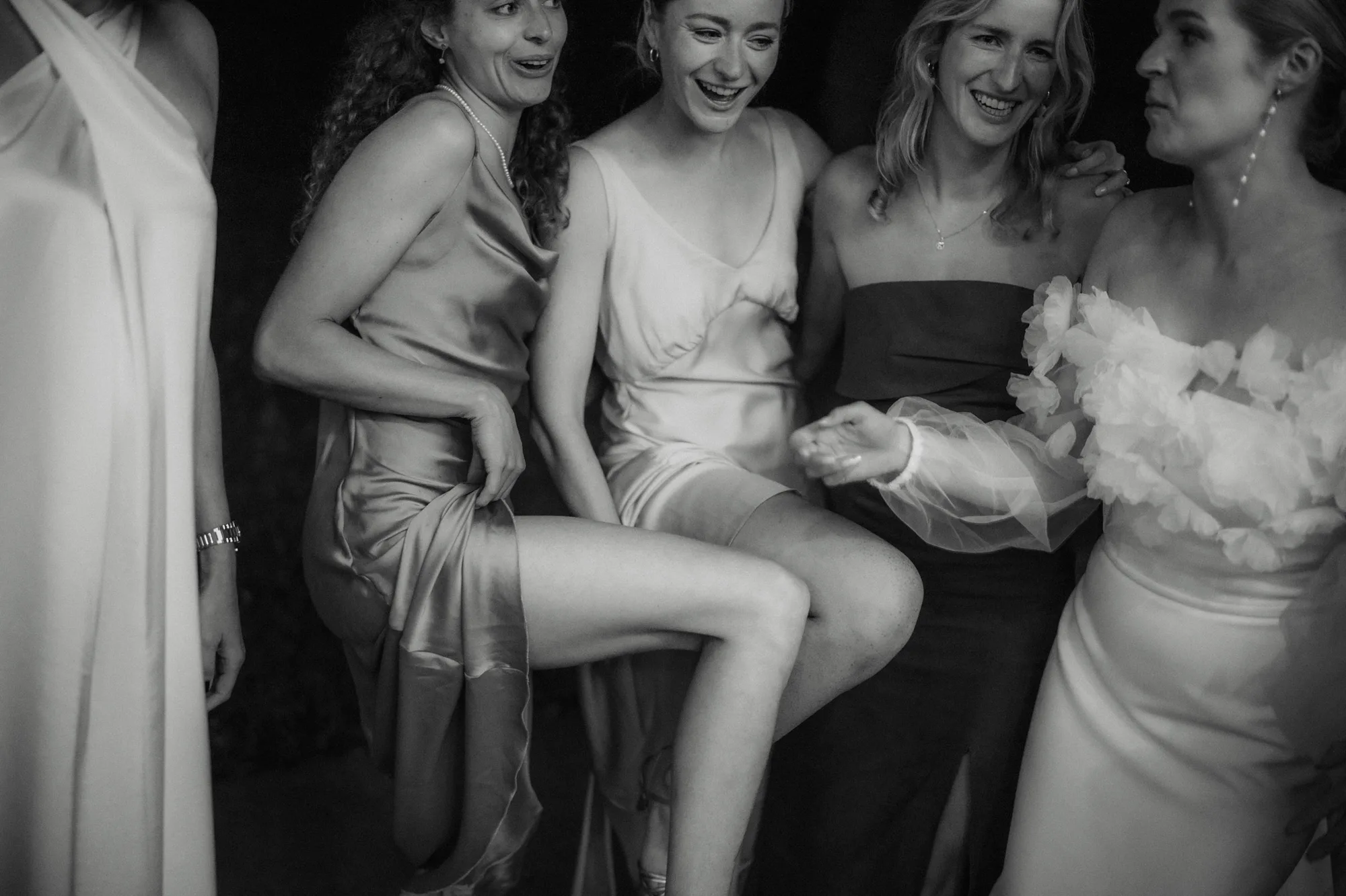 Five women dressed in elegant evening gowns smiling and interacting joyfully, one lifting her leg playfully.