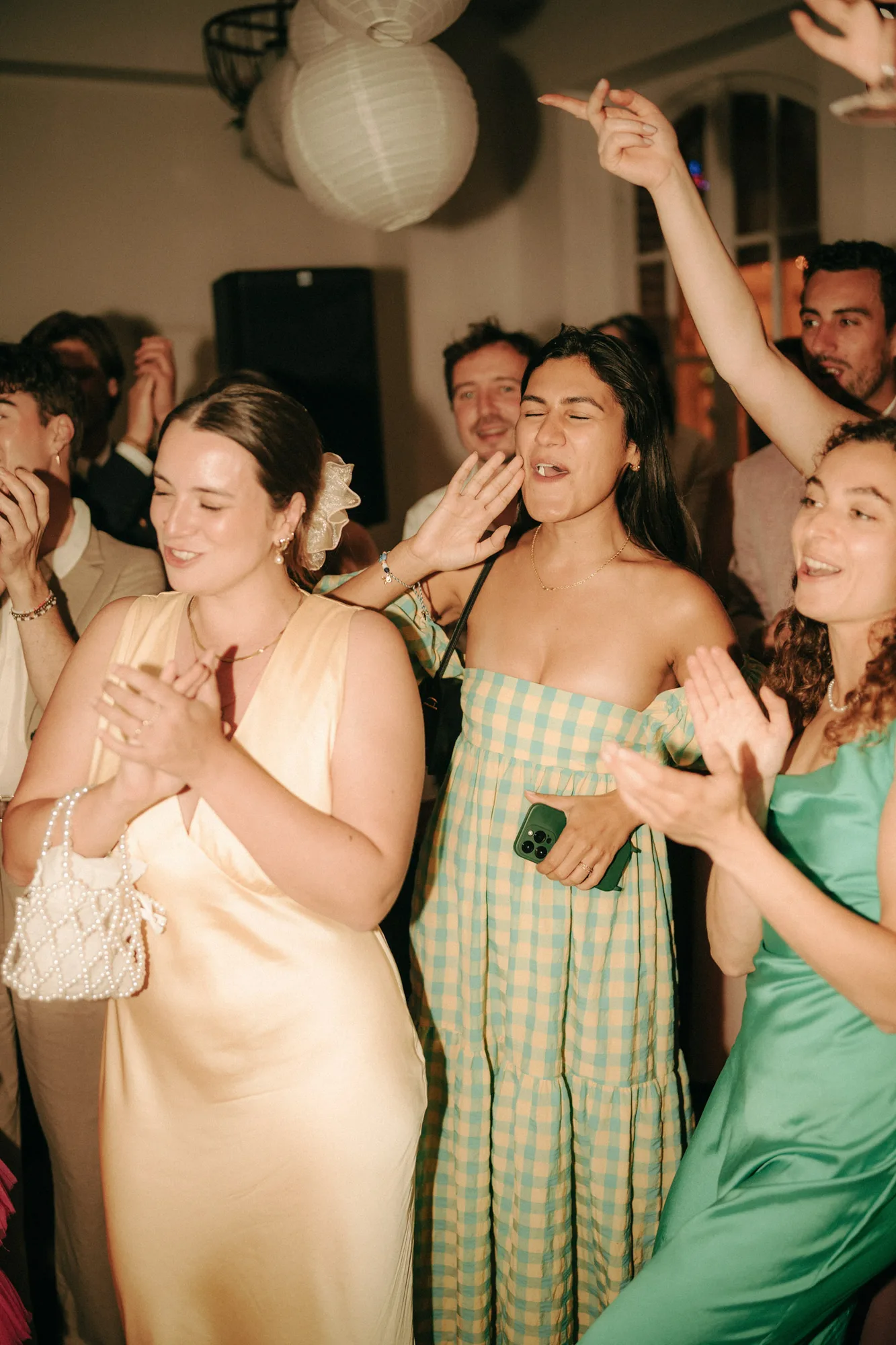 Group of people joyfully singing and clapping at a party, with three women prominently in colorful dresses.