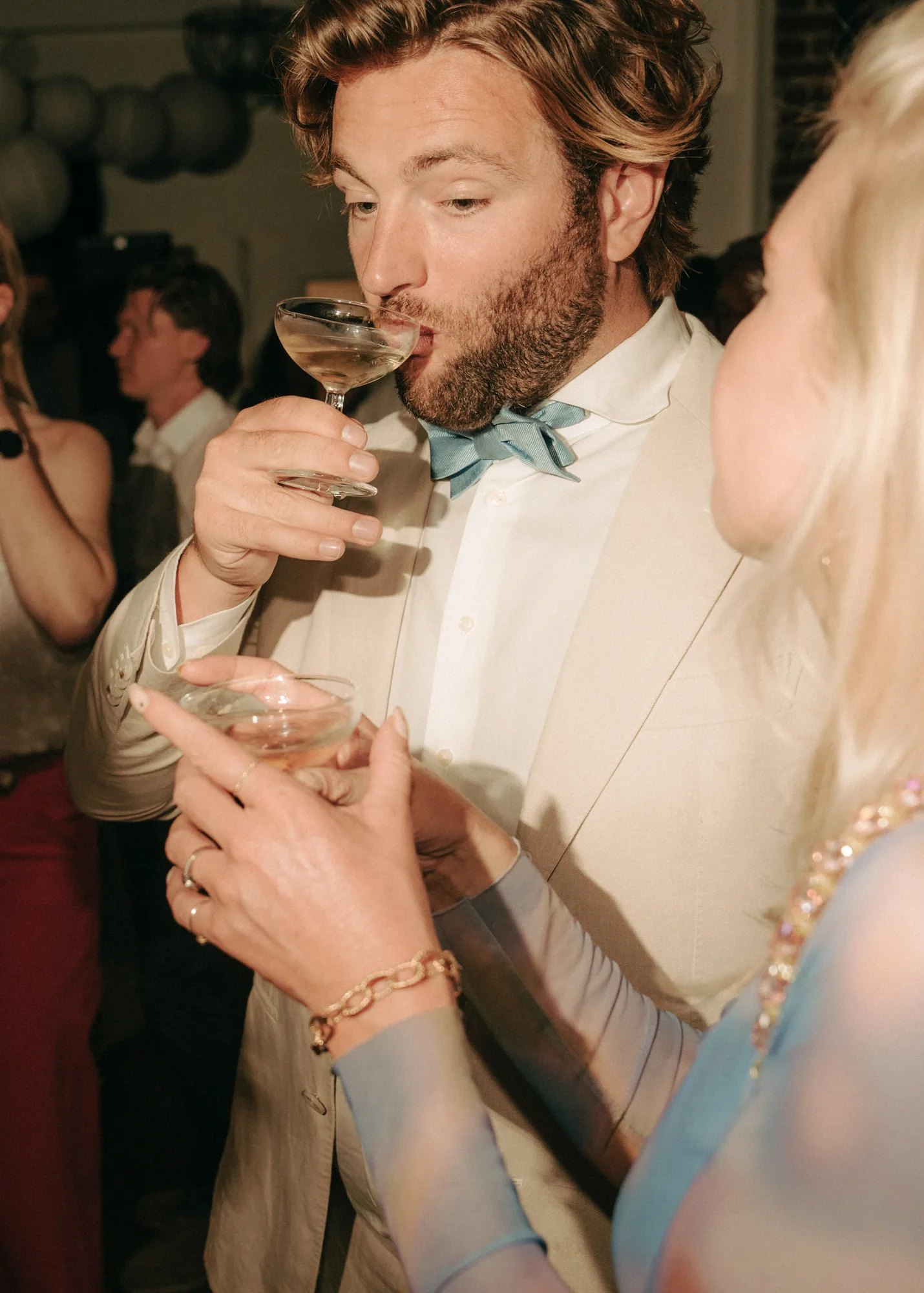 Bearded man in a light suit and bow tie drinking from a cocktail glass while a woman holds another glass nearby at a social event.