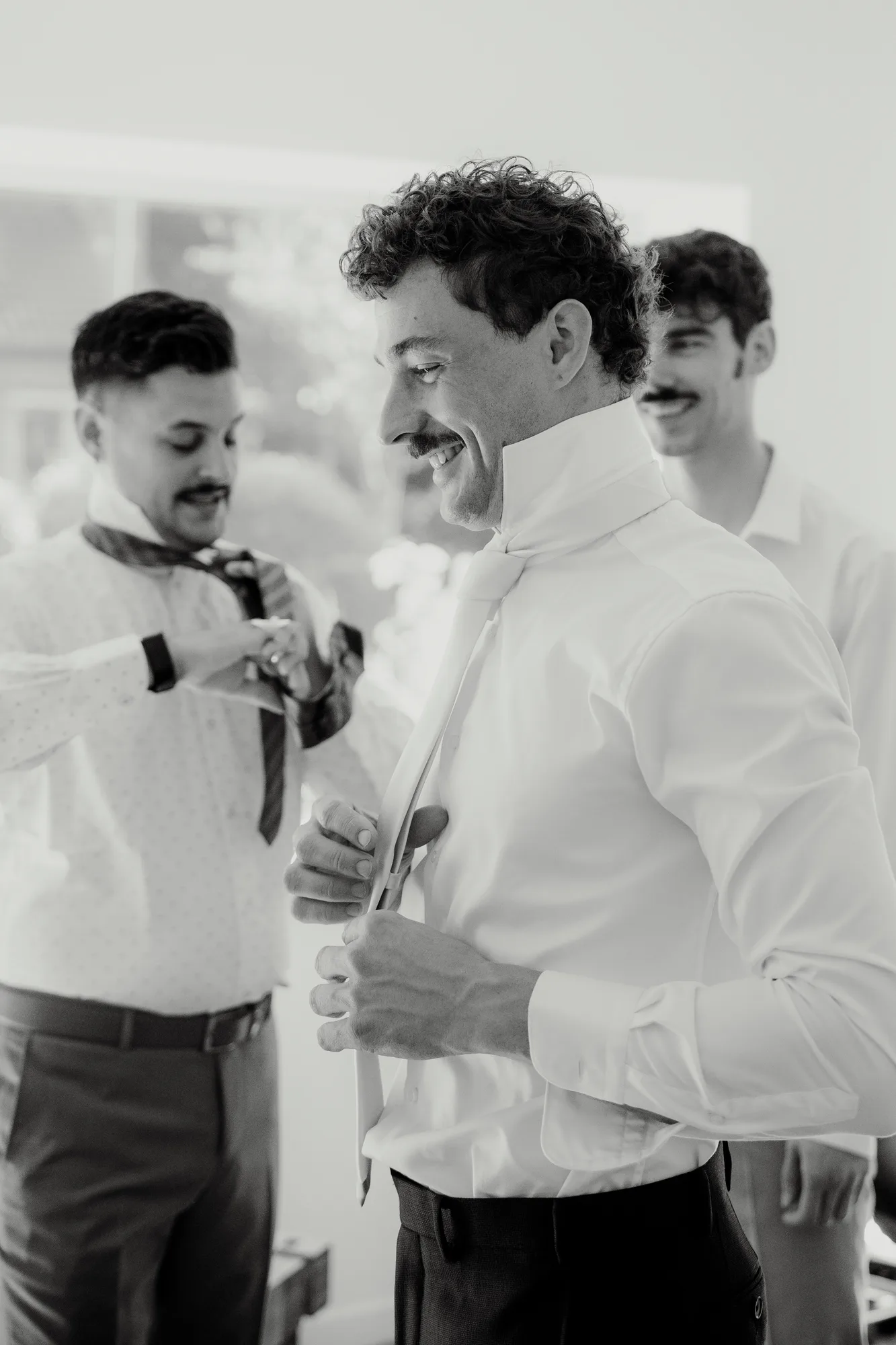 Three men smiling indoors while tying their neckties, preparing for a formal event.