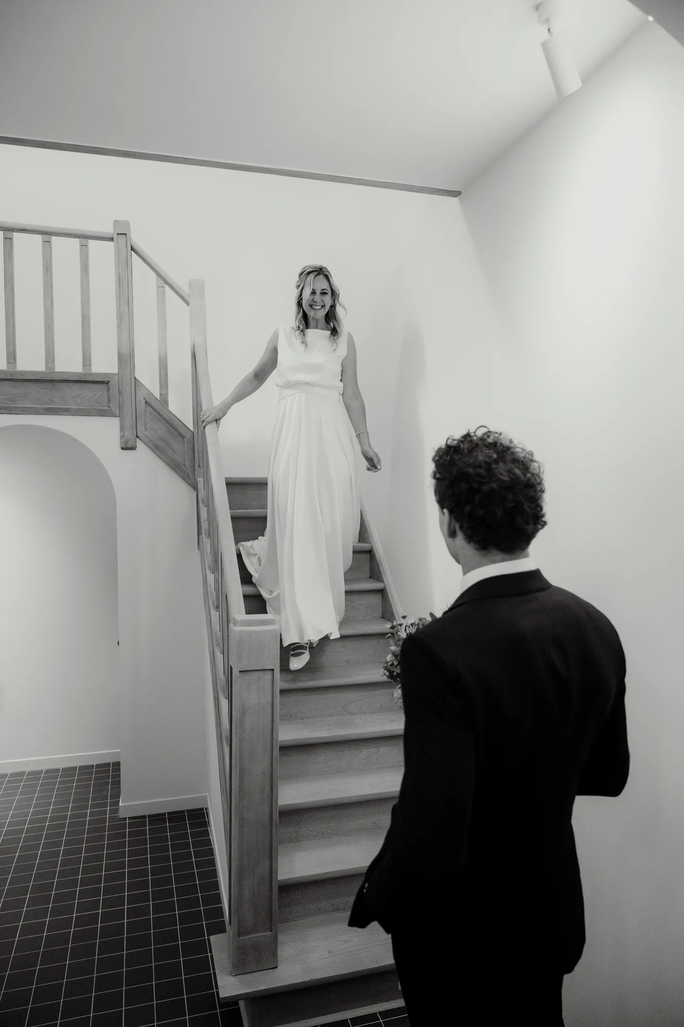 Smiling woman in a white dress descending wooden stairs toward a man in a suit holding a bouquet.