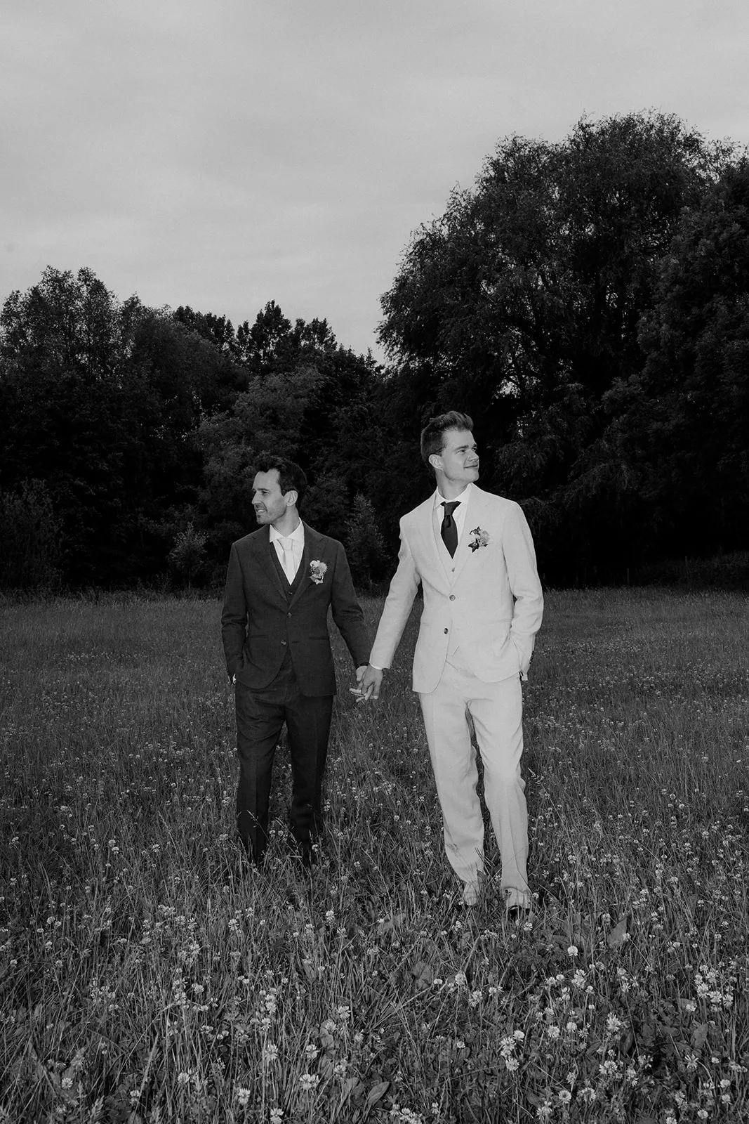 Two grooms in suits holding hands and walking in a flower-filled field with trees in the background.