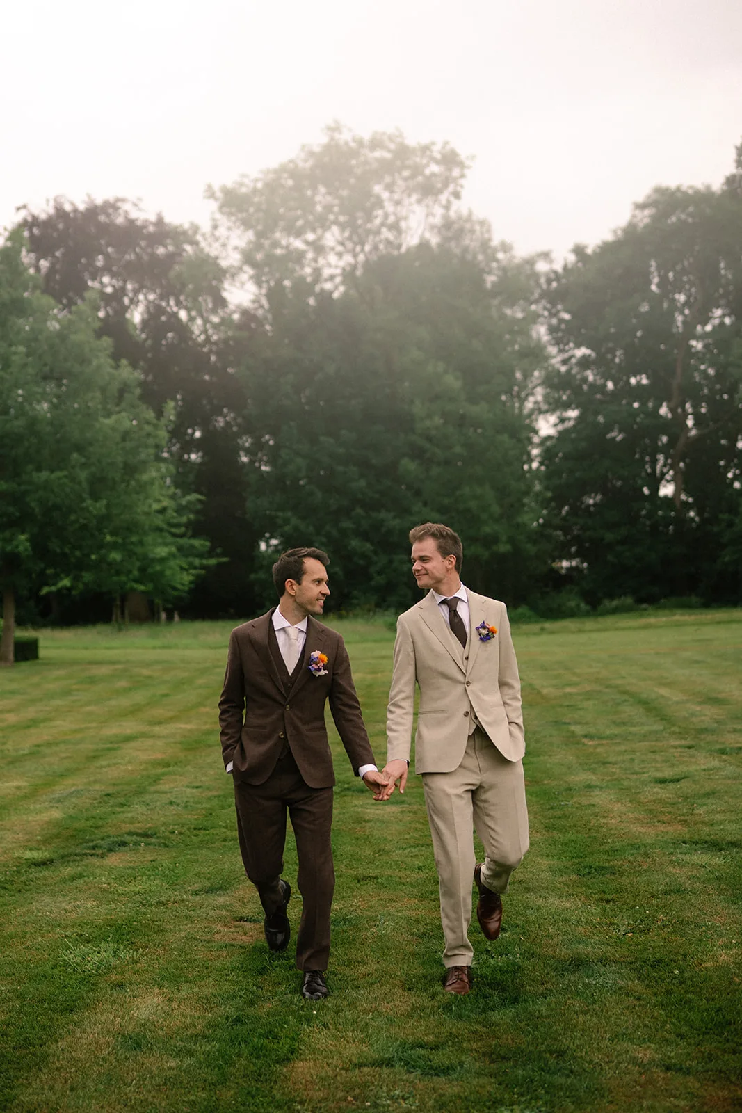 Two men in suits holding hands and walking on green grass in a park with trees in the background.