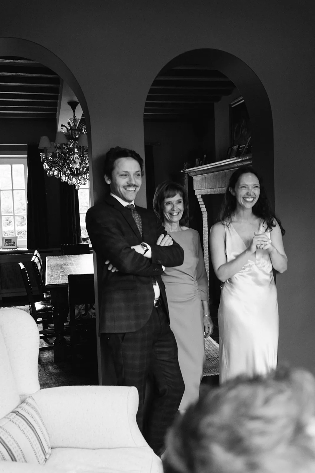 Three people smiling and standing in an indoor setting, with one man in a suit and two women in dresses near an archway.