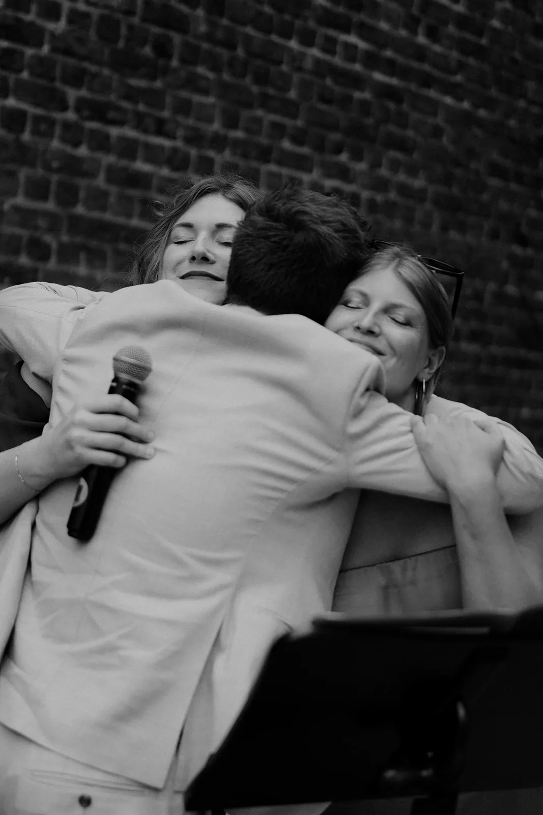 Black and white photo of three people sharing a warm group hug, two women smiling with eyes closed, one holding a microphone.