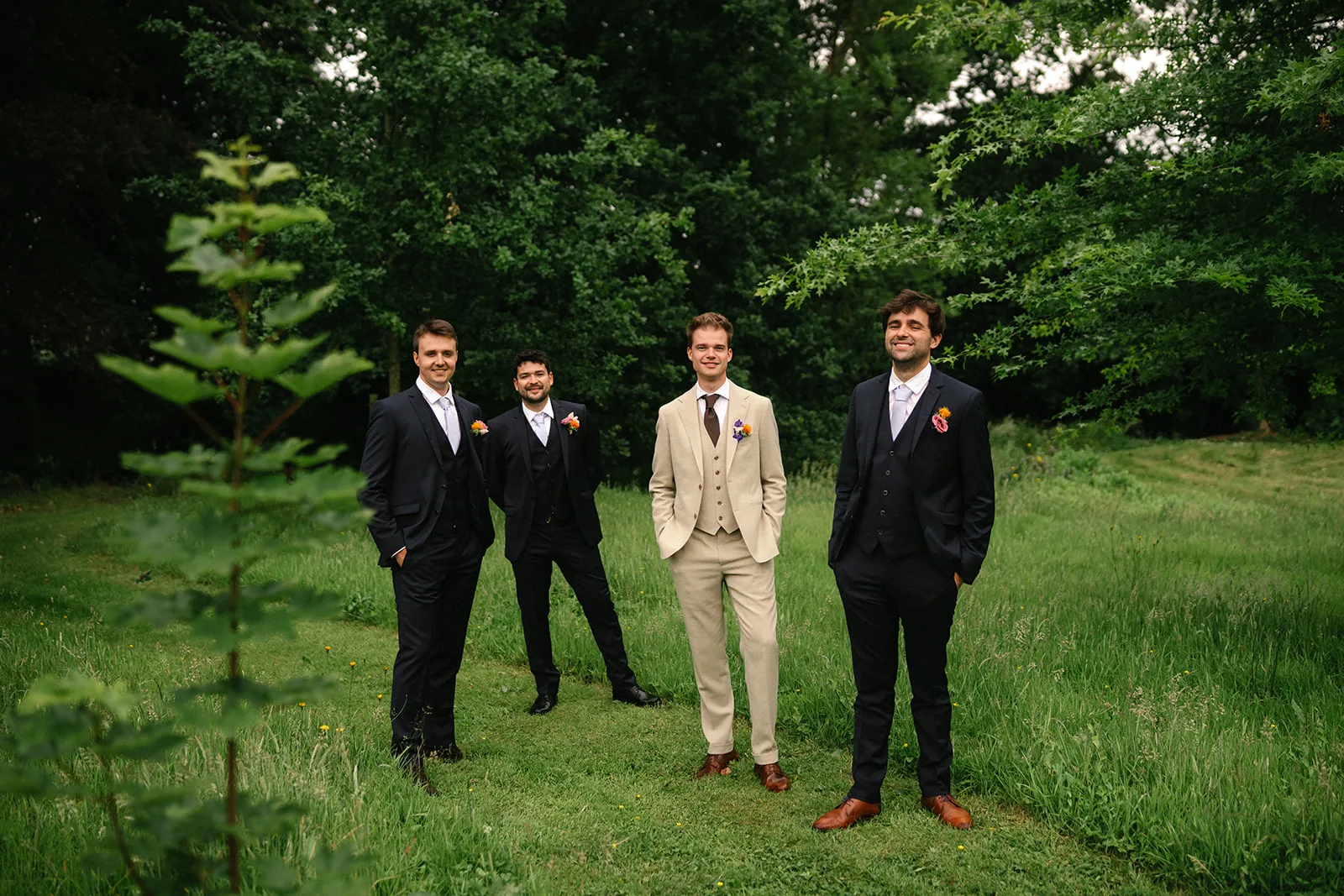 Four men in suits standing on grass in a wooded area, with one man wearing a light beige suit and the other three in dark suits.