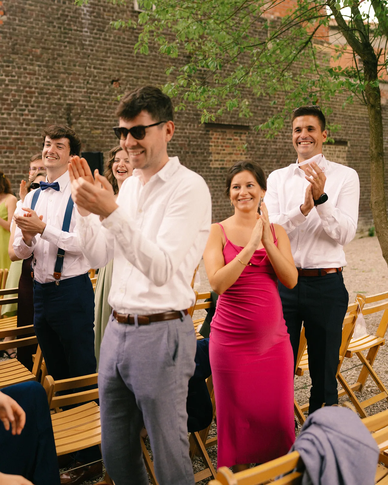 Group of people standing outdoors, smiling and clapping in celebration at an event with wooden chairs and a brick wall background.