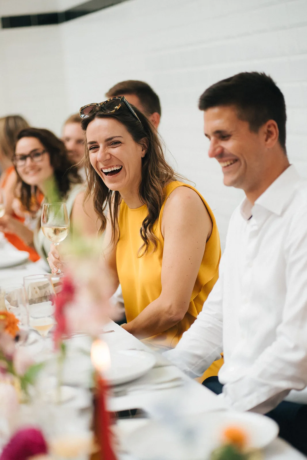 Group of people sitting at a table, a woman in a yellow dress holding a glass of white wine and laughing with others smiling around her.