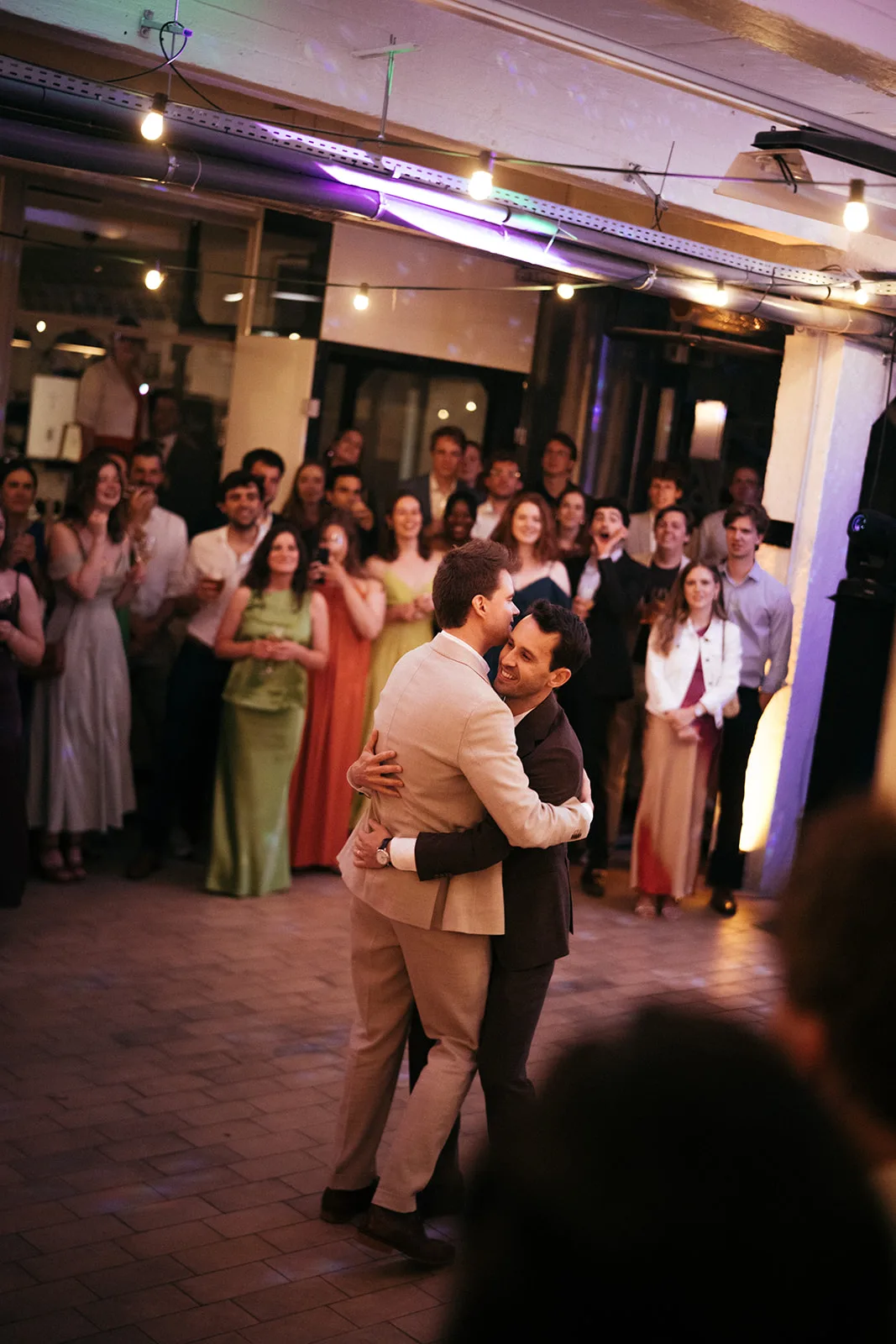 Two men in suits warmly embracing and dancing at a celebration with a crowd of smiling guests watching in the background.
