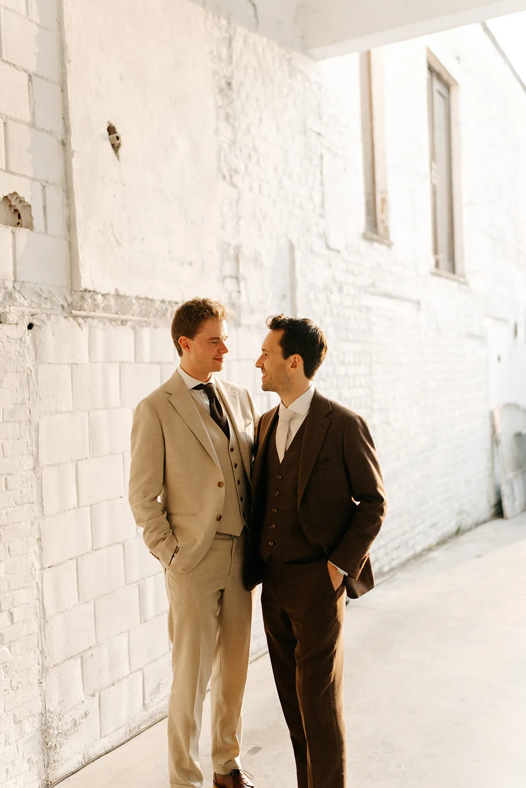 Two men dressed in suits standing close together and smiling at each other against a white brick wall.