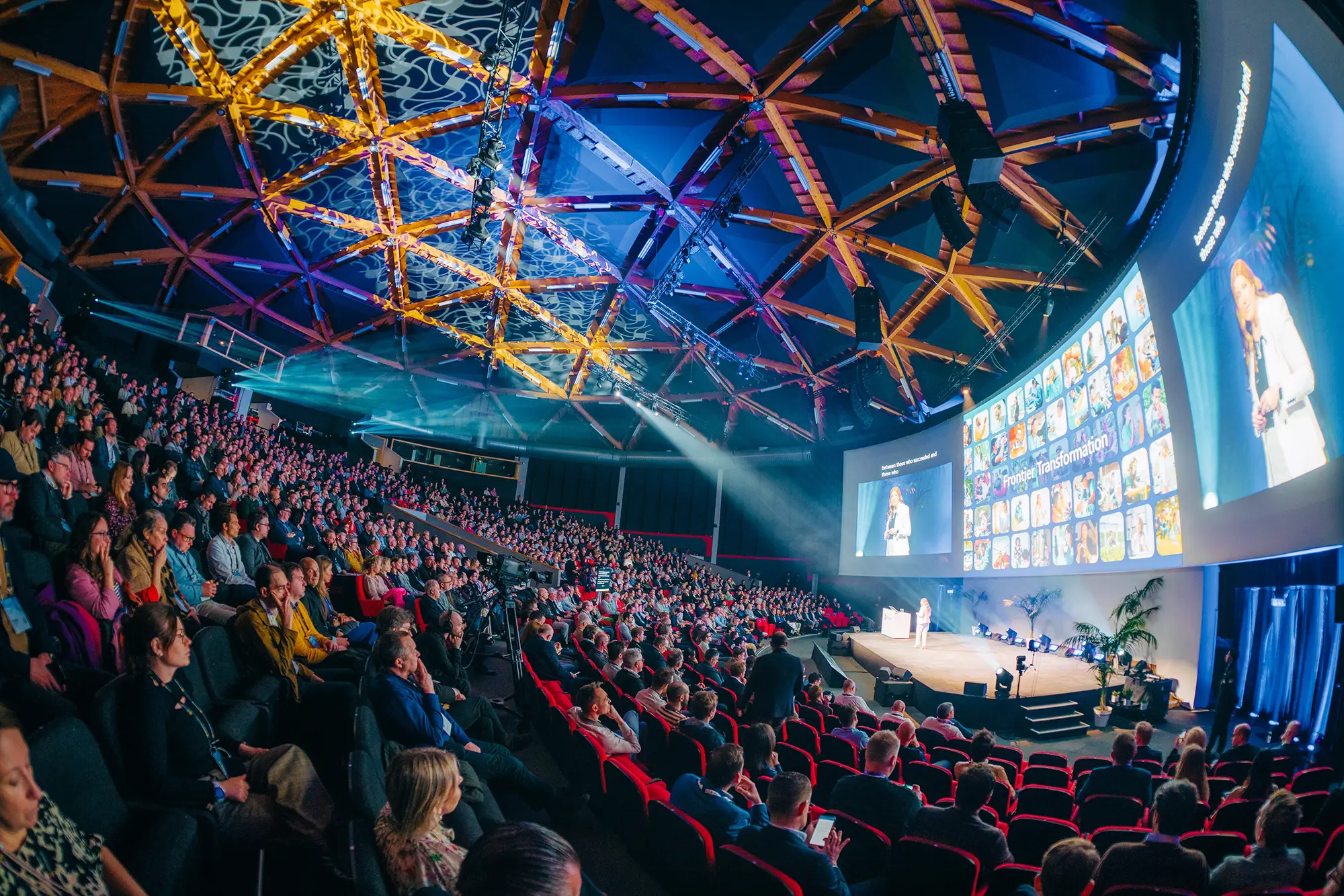 Large auditorium with a crowd watching a female speaker on stage presenting in front of a curved screen displaying a 'Frontier Transformation' slide.