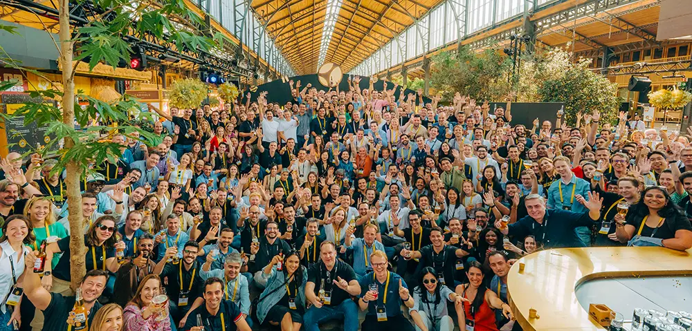 Large group of people at an indoor event space, smiling and waving toward the camera under a high glass ceiling.