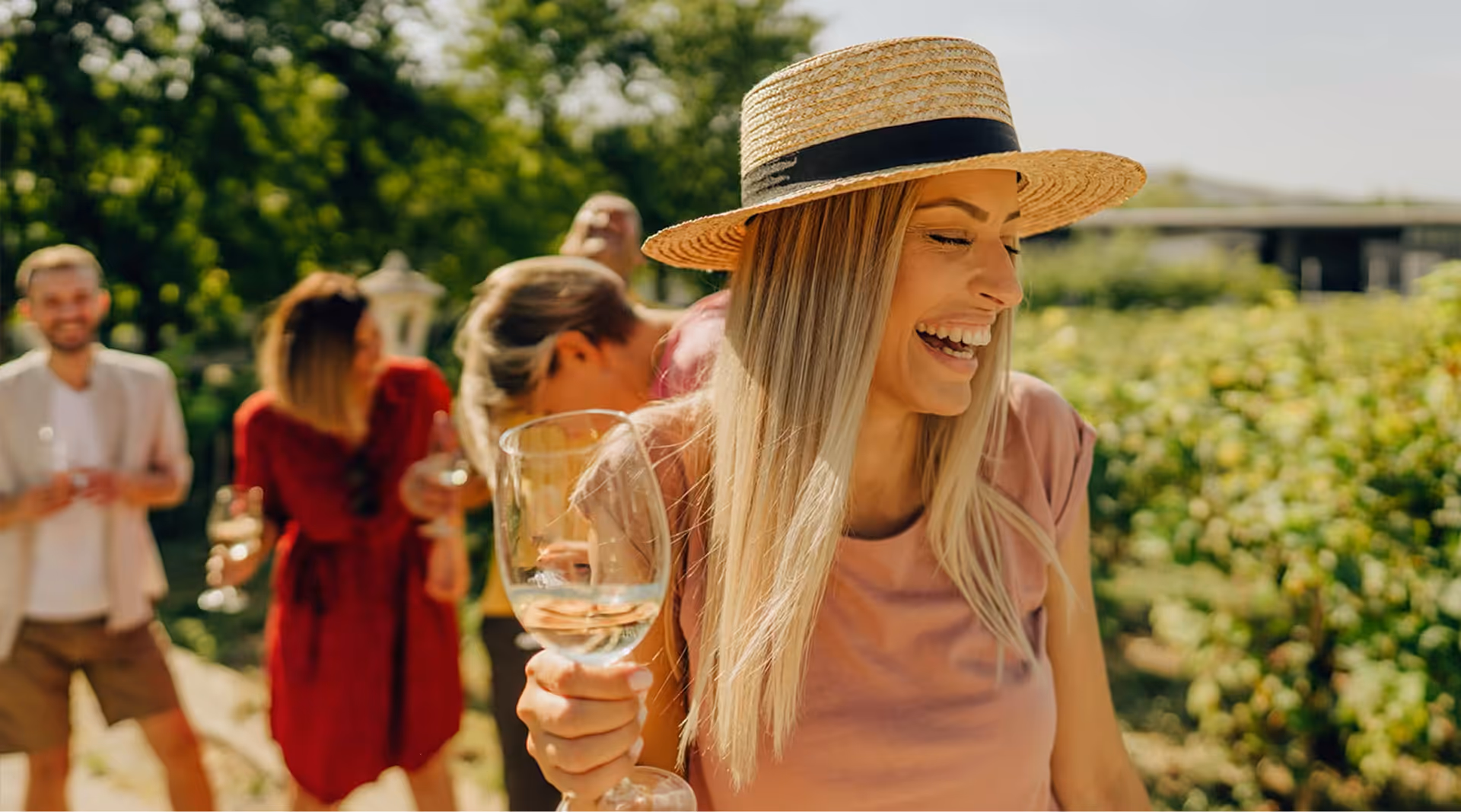 A smiling young woman in a straw hat holding a glass of white wine during an outdoor vineyard tasting experience at The Swift Water hotel.