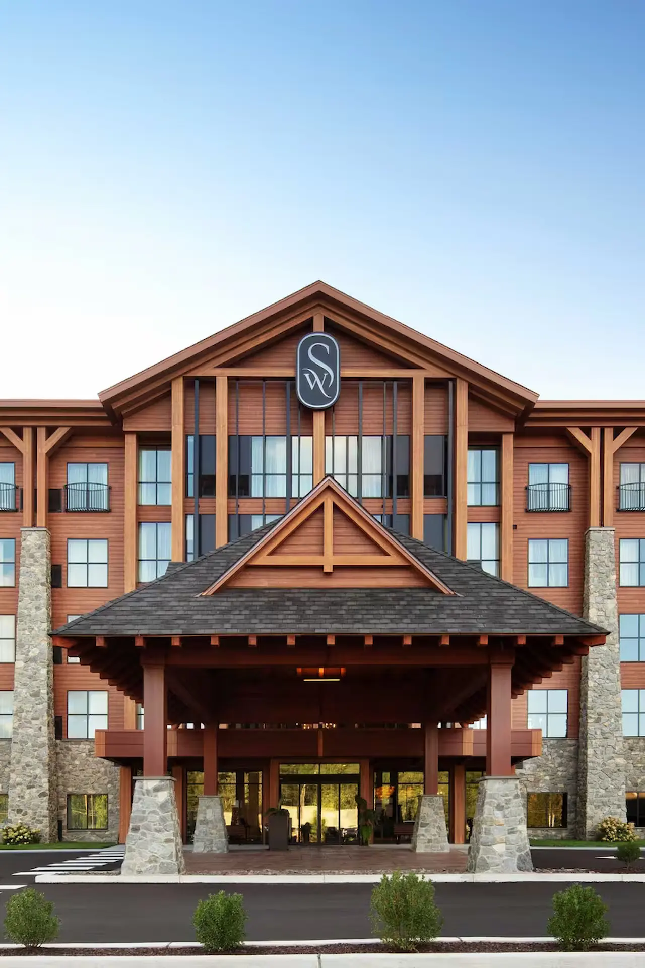 Panoramic view of The Swift Water hotel's grand lobby featuring high wood-paneled ceilings, a sparkling chandelier, stone walls, and a modern reception desk.