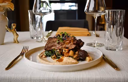 An appetizing close-up photograph of the main course at The Swiftwater hôtel, featuring braised short ribs on a bed of creamy mashed potatoes and sautéed kale, elegantly presented on a white plate with gold cutlery.