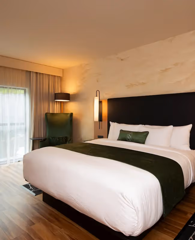 A spacious and bright guest room at The Swiftwater hotel, featuring a plush King bed with crisp white linens, a forest green bed runner, and a stylish armchair by the window.