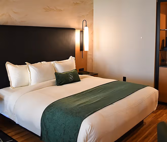 Close-up of a luxurious bed at The Swiftwater with crisp white linens, a dark headboard, and a signature green velvet lumbar pillow against a decorative wall.