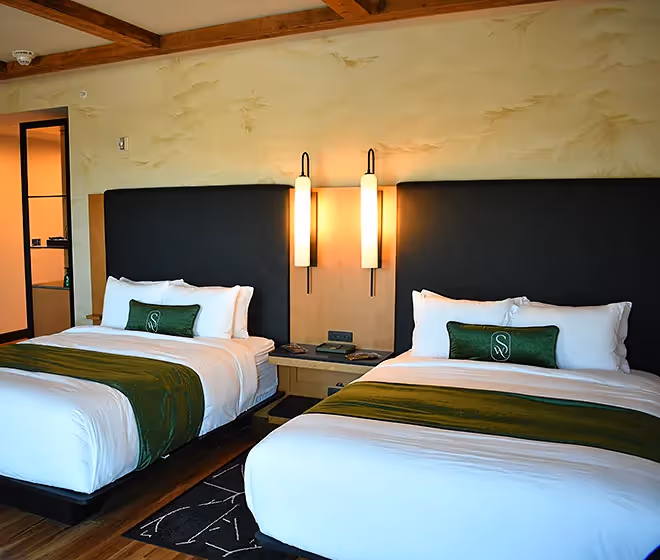An elegant hotel room at The Swiftwater featuring two double beds with emerald green runners, modern black headboards, and warm ambient lighting.