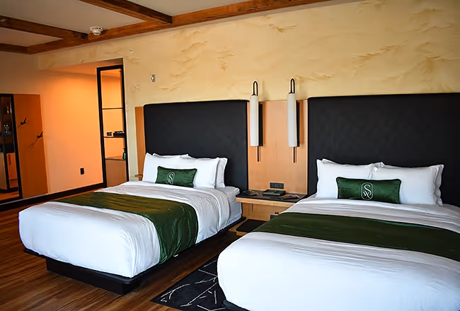 Close-up of two queen beds at The Swiftwater hotel with crisp white linens, emerald green throw pillows, and warm ambient lighting.