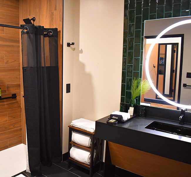 Contemporary bathroom at The Swiftwater featuring a black vanity with a circular backlit mirror, wood-textured shower tiles, and matte black fixtures.