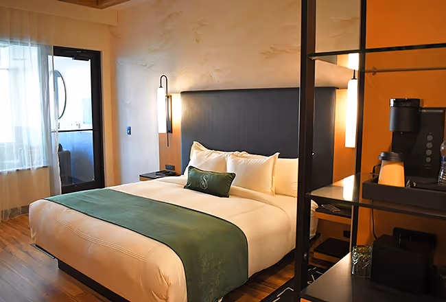 A luxury guest room at The Swiftwater hotel featuring a plush king-sized bed with a green runner, modern wall sconces, and a view into a private spa area.