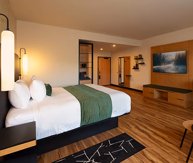 A stylish hotel room at The Swiftwater featuring a king bed with forest green accents, warm wood flooring, and a large wall-mounted TV.