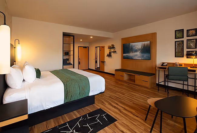 A stylish hotel room at The Swiftwater featuring a king bed with forest green accents, warm wood flooring, and a large wall-mounted TV.