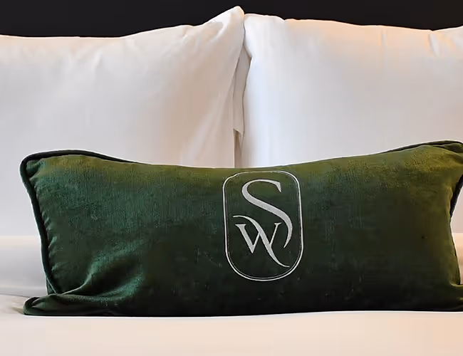 An emerald green velvet lumbar pillow featuring an embroidered "SW" logo, resting on a luxurious white bed at The Swiftwater hotel.