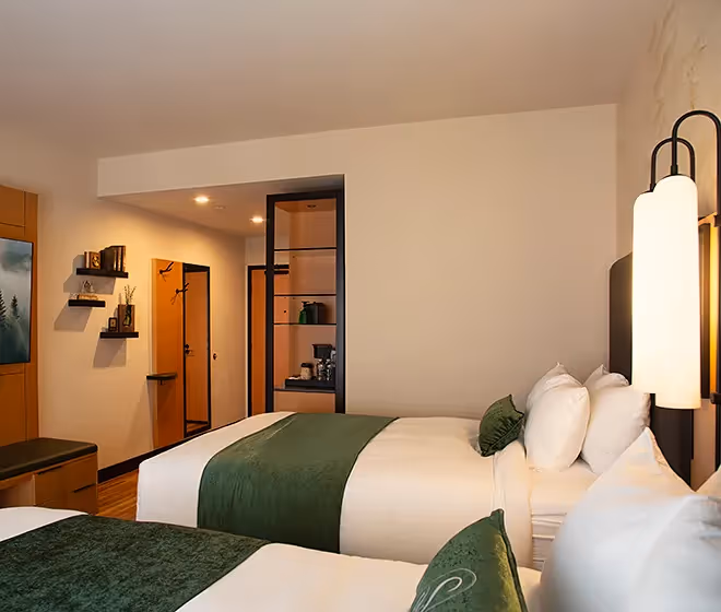 An elegant guest room at The Swiftwater hotel featuring two double beds with forest green runners and warm ambient lighting.