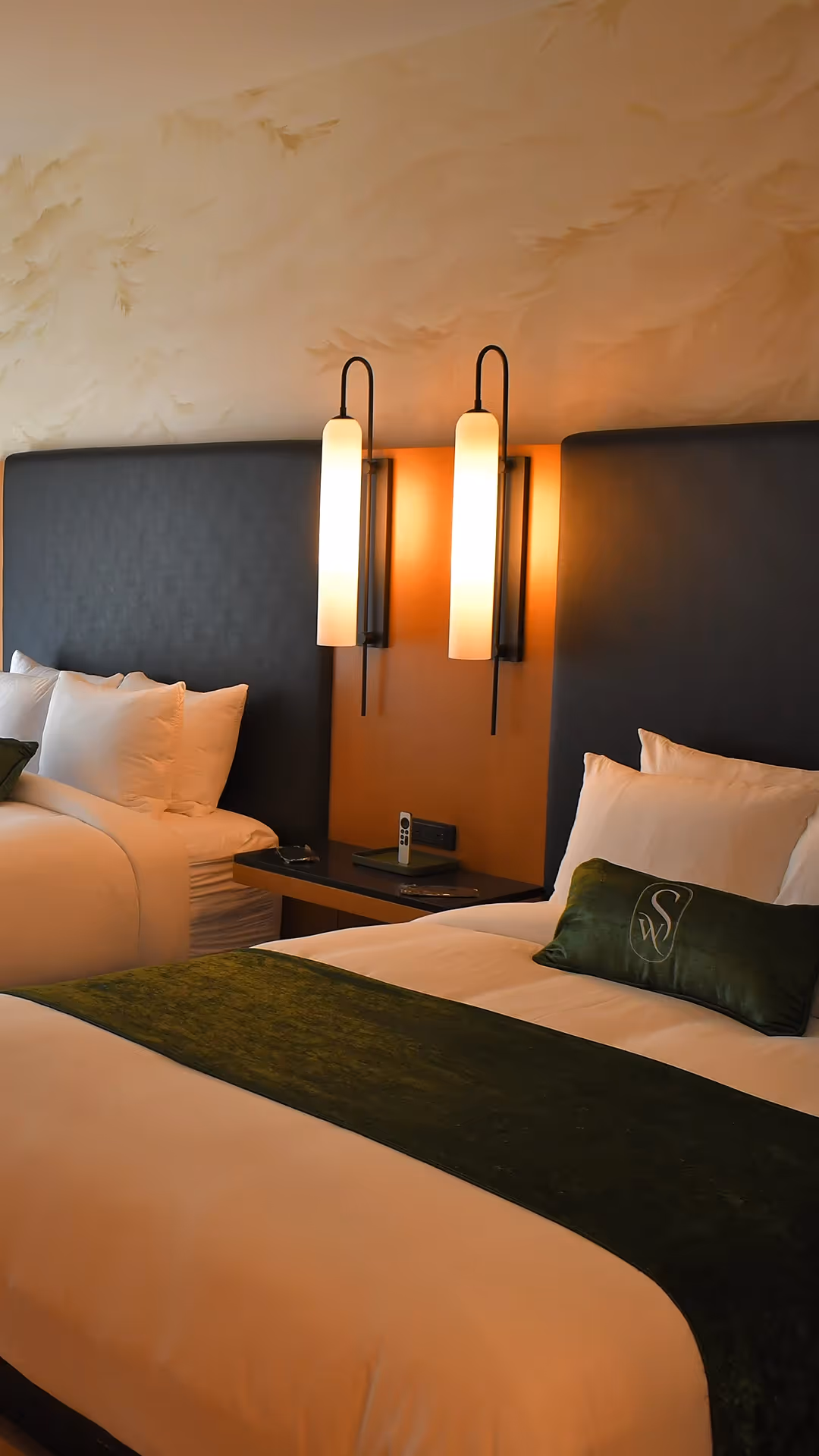 Guest room at The Swiftwater hotel featuring two beds, white linens, forest green accents, and warm lighting.