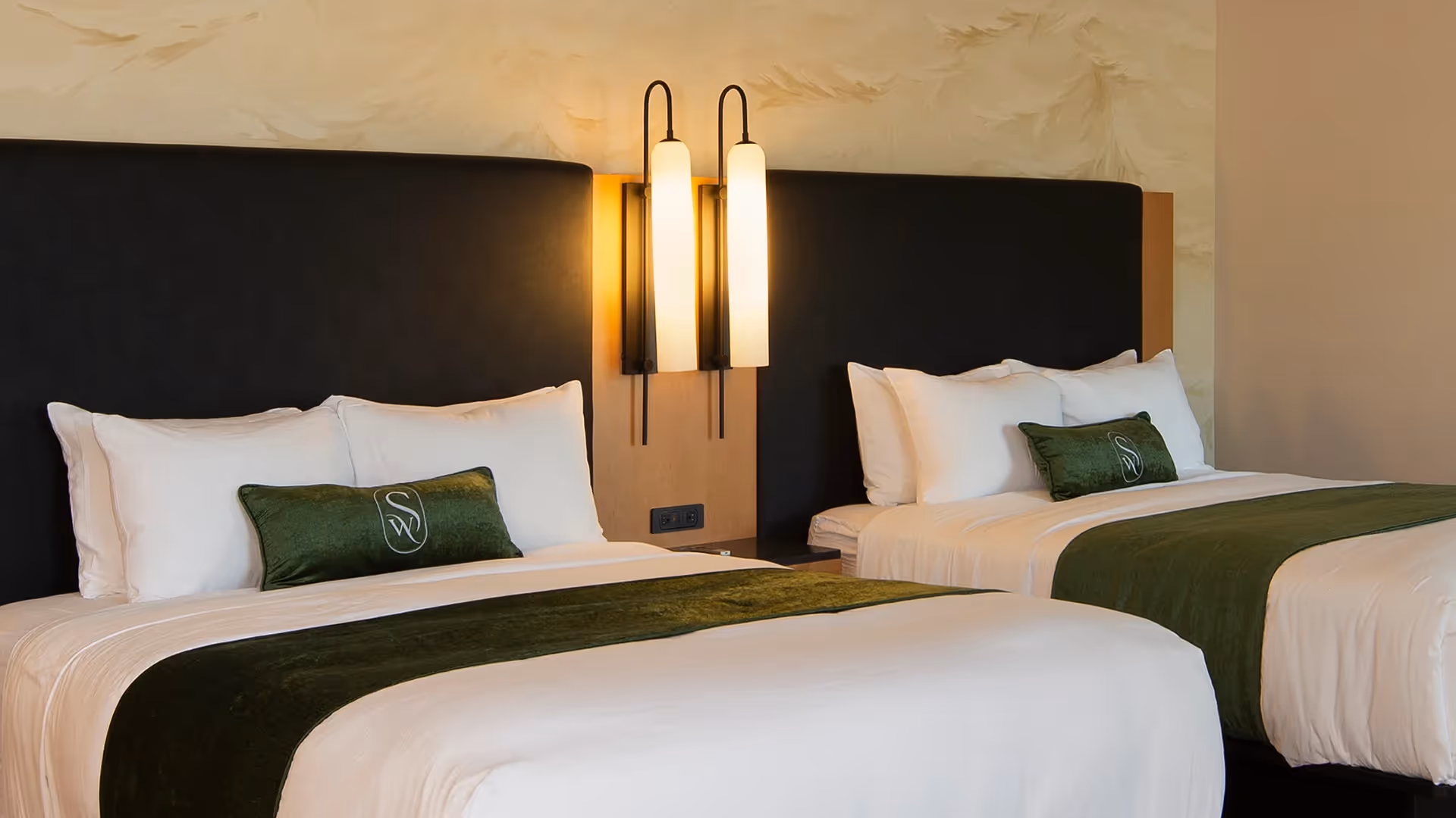 Close-up of two queen beds at The Swiftwater hotel with crisp white linens, emerald green throw pillows, and warm ambient lighting.