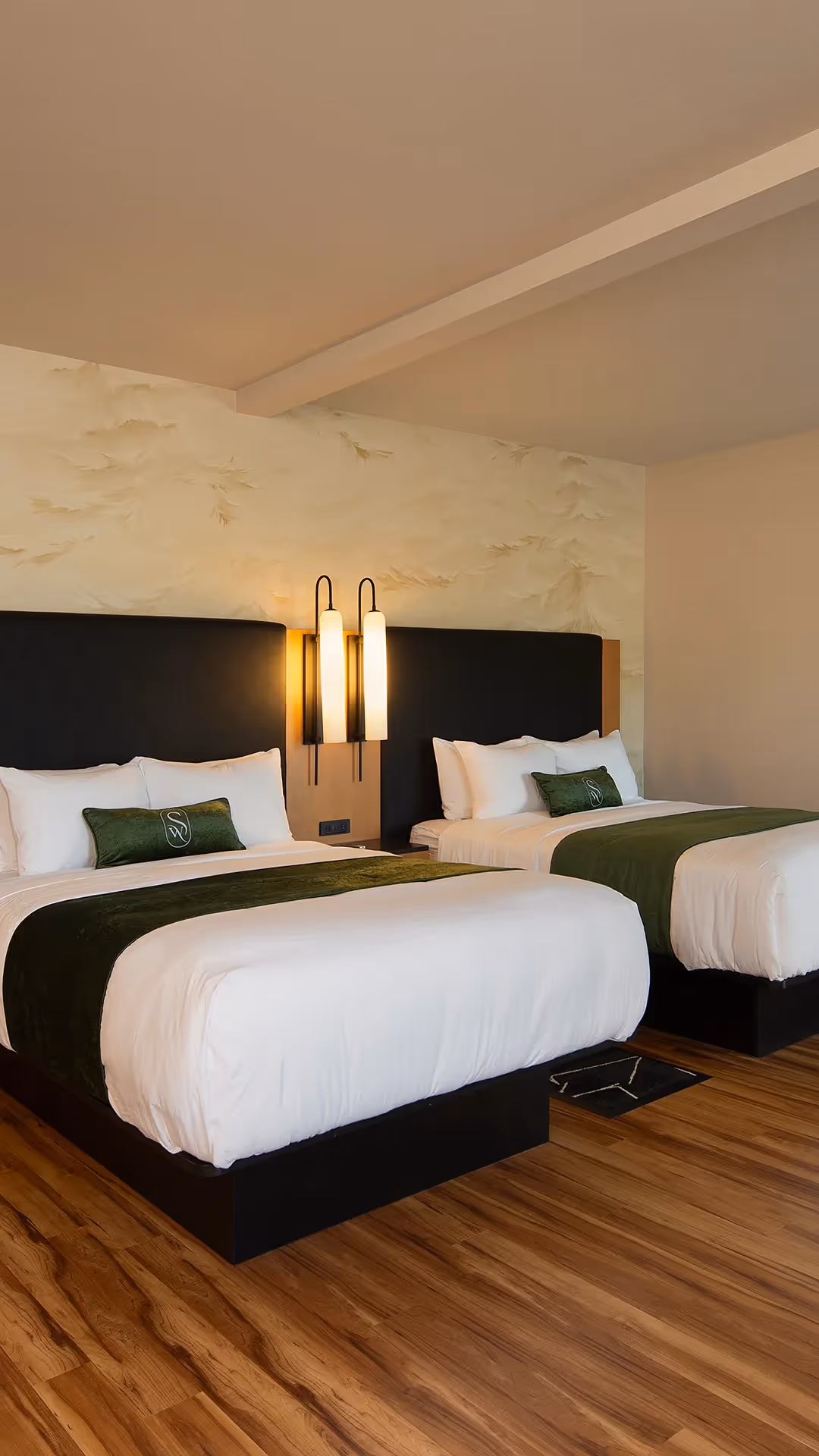 Close-up of two queen beds at The Swiftwater hotel with crisp white linens, emerald green throw pillows, and warm ambient lighting.