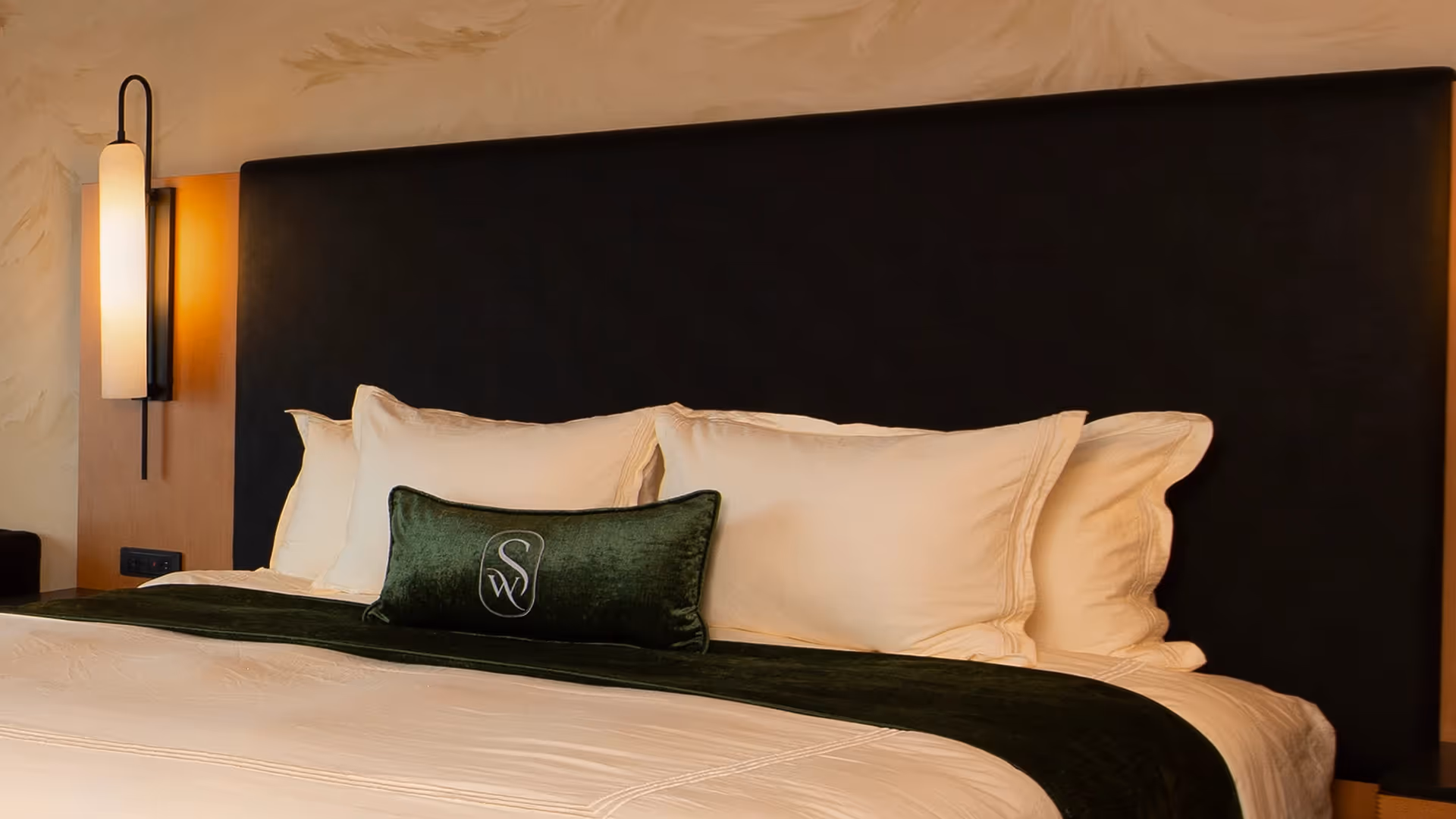 A welcoming close-up of the luxurious bed at The Swiftwater hotel, featuring plush white pillows, an emerald green accent throw, and a sleek dark headboard.