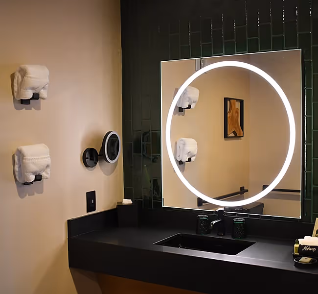 Chic vanity area at The Swiftwater hotel, featuring a glowing circular LED mirror and a sleek black counter with premium toiletries.